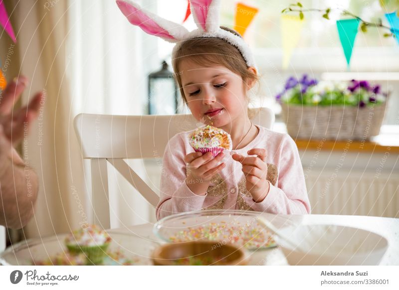 Vater und Tochter feiern Ostern und essen mit Glasur überzogene Törtchen. Fröhlicher Familienurlaub. Süßes kleines Mädchen in Hasenohren. Wunderschön dekorierter Raum