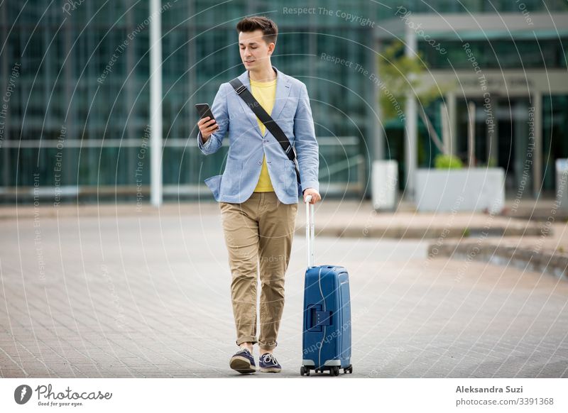 Stylischer Mann steht am Flughafen mit Smartphone und Koffer, blättert, textet, benutzt mobile Apps. Geschäftsreisende. Anwendung Ankunft Tasche Business