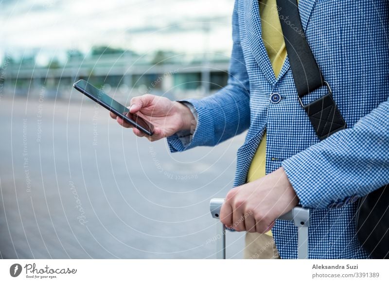Stylischer Mann steht am Flughafen mit Smartphone und Koffer, blättert, textet, benutzt mobile Apps. Geschäftsreisende. Anwendung Ankunft Tasche Business
