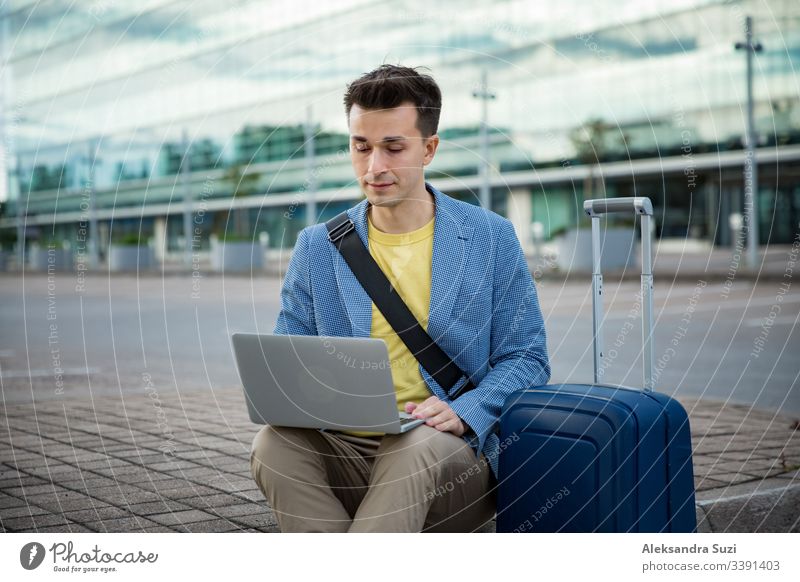 Ein stilvoller Mann sitzt am Flughafen mit Koffer und Laptop, arbeitet, tippt, stöbert. Geschäftsmann auf Reisen. Erwachsener Tasche Gepäck Browsen Business