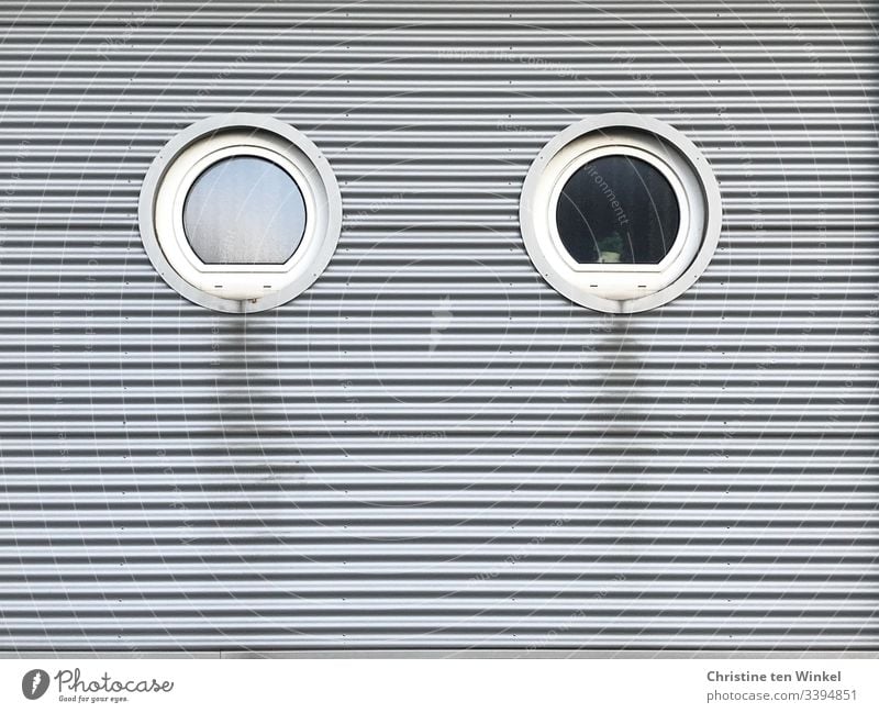 Zwei runde Fenster in einer verkleideten Hausfassade weiß Fassade Gebäudefassade schmutzig silber Fassadenverkleidung silberfarben grau trist Streifen Linien