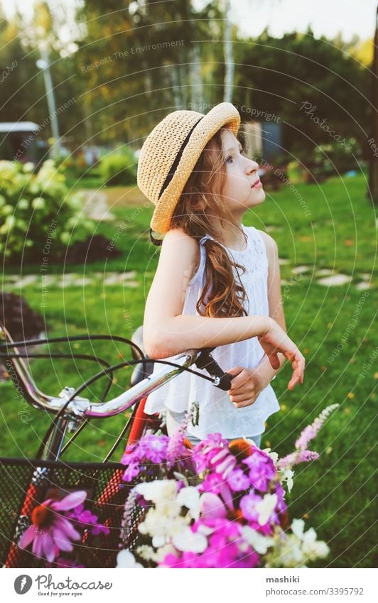 fröhliches Kind Mädchen mit Hut Fahrrad fahren mit Wildblumenstrauss, Sommerferienkonzept Buch Kindheit im Freien Natur Park Spaß Glück Lifestyle lesen Freizeit