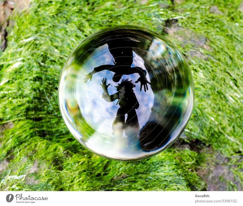Die Silhouetten von Mann und Frau werden in einer Glaskugellinse, die auf dem grünen Gras liegt, verzerrt reflektiert. Algen Ball blau Wolken gekrümmter Spiegel