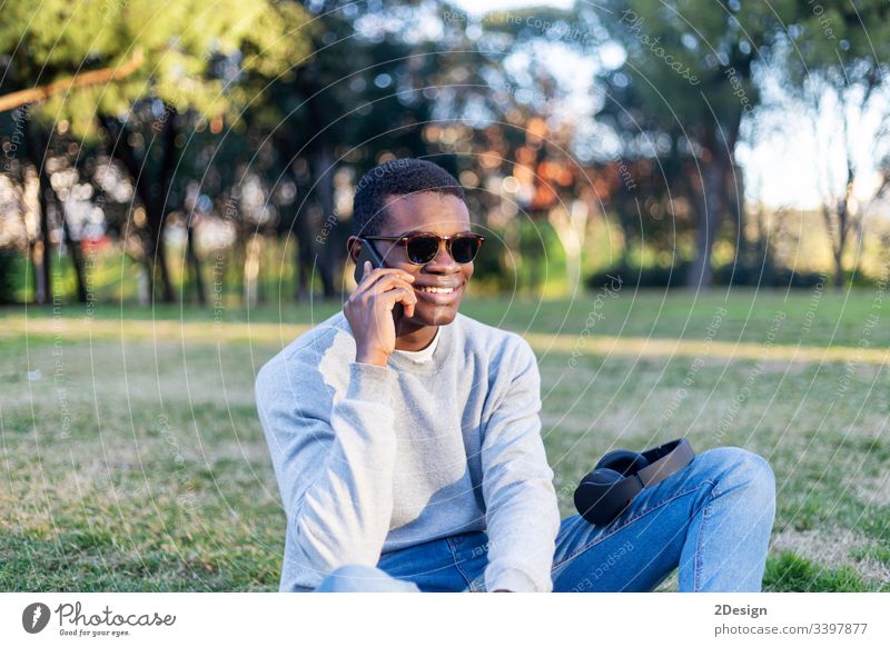 Modischer schwarzer Mann mit Sonnenbrille, der im Freien sitzt und seinen Nachrichten-Feed oder seine Nachrichten über soziale Netzwerke per Handy abruft