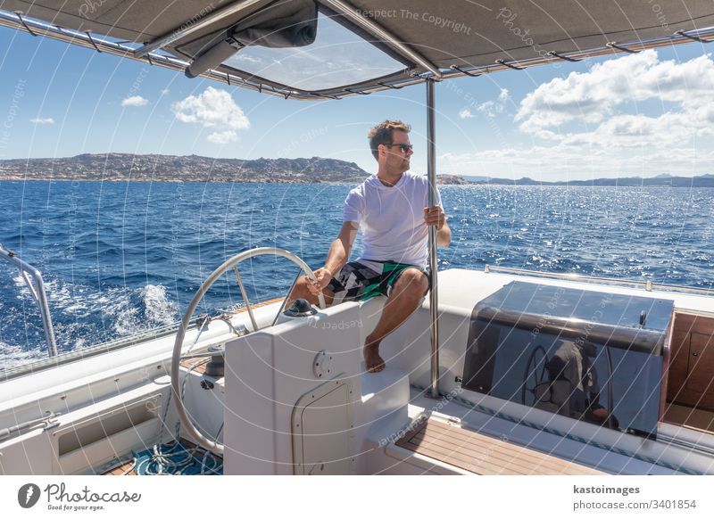 Attraktiver männlicher Skipper, der das schicke Katamaran-Segelboot an einem sonnigen Sommertag auf ruhigem blauen Meerwasser steuert. nautisch Lenkrad Boot