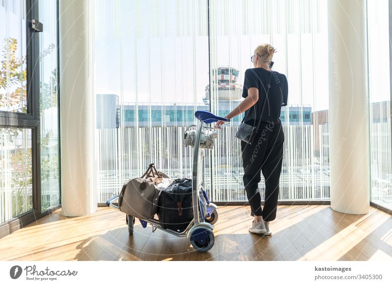 Lässige Frau am Flughafenterminal, die am Handy telefoniert und auf den Abflug des Fluges mit Gepäck auf dem Gepäckwagen wartet. reisen Karre Abheben Telefon