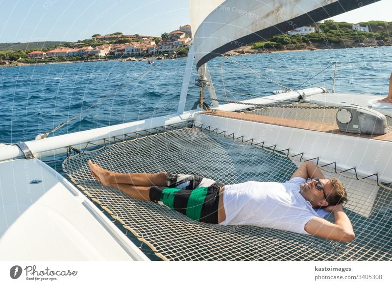 Sportlicher Mann entspannt sich, in der Hängematte eines Katamaran-Segelbootes liegend, auf luxuriösen nautischen Ferien in der Nähe der bildschönen Stadt Palau, Sardinien, Italien.