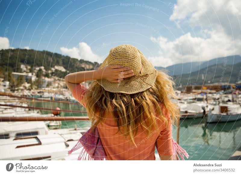 Porträt einer Frau mit leicht gelocktem Haar, Rückansicht, mit der Hand Strohhut haltend. Sonniger Hafen, Boote und Jachten, grüne Berge im Hintergrund. Das Leben genießen, glücklich reisen,