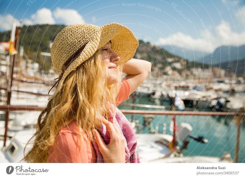 Porträt einer jungen Frau mit hellem lockigem Haar und Strohhut, die lächelnd Sonne und Wind genießt. Sonniger Hafen mit Booten und Jachten, grüne Berge im Hintergrund. Lebensfreude, fröhliche Reisende,