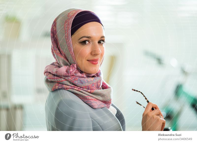 Schöne, stilvolle Frau mit Hijab und Brille, die im Büro am Schreibtisch mit Laptop sitzt. Porträt einer selbstbewussten muslimischen Geschäftsfrau. Modernes Büro mit großem Fenster, Fahrrad im Hintergrund.