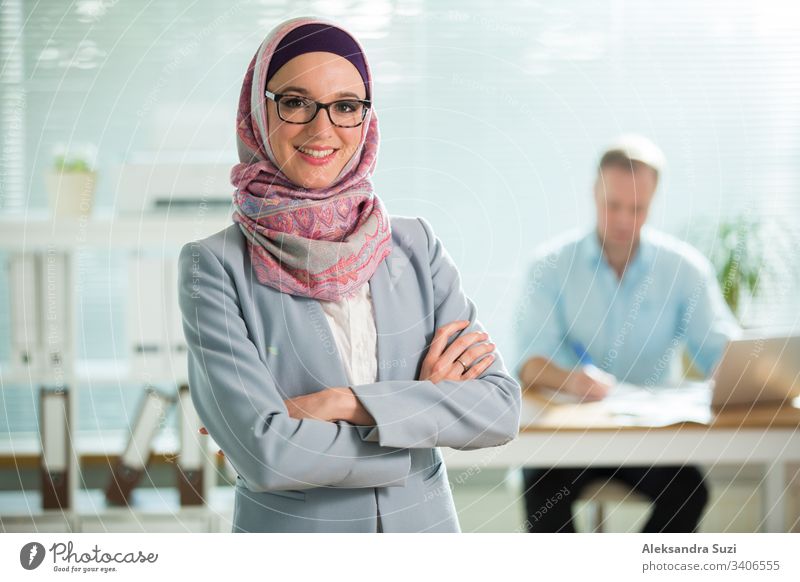 Hübsche junge Arbeiterin in Hijab, Anzug und Brille steht im Büro und lächelt. Porträt einer selbstbewussten muslimischen Geschäftsfrau. Modernes Büro mit großem Fenster, im Hintergrund ein Mann am Schreibtisch arbeitend.