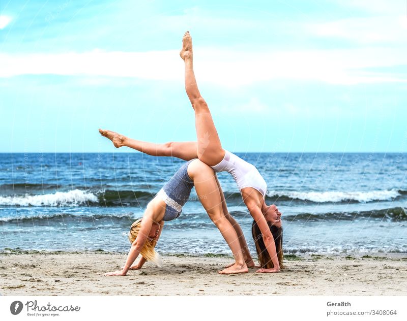 zwei junge Mädchen in weißem Oberteil und schwarzem Unterteil am Sandufer akrobatisch Akrobatisches Yoga aktiv Asana Strand blond blau Körper braunhaarig