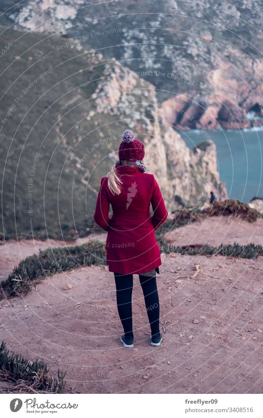 Junge Frau in rotem Mantel, die von der Spitze einer Klippe das Meer betrachtet Stehen atlantisch Wasser malerisch Wahrzeichen Saum Marin Licht Raum Sintra