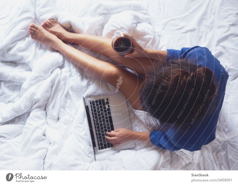 Junge Frau trinkt zu Hause in ihrem Bett Kaffee und schaut auf ihren Laptop gemütlich Morgen Tasse Mädchen heimwärts Buch Beine weich Top Ansicht jung warm