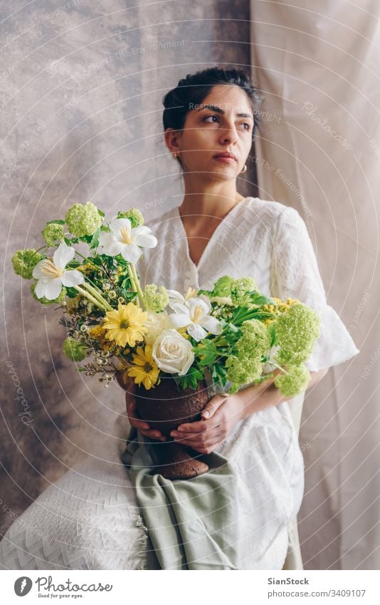 Junge Frau in einem weißen Kleid, die eine Vase mit Blumen hält. Vintage, romantisches Konzept. Blumenstrauß Mädchen Metall sitzen Sitzen Stuhl weiches Licht