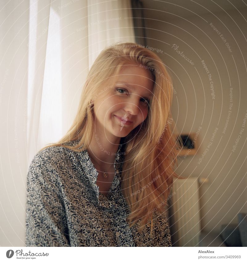 Portrait einer jungen Frau neben dem Fenster an der Gardine Mädchen blond schön schlank anmutig elegant lifestyle Wohnen Wohnung zu Hause Bluse Muster