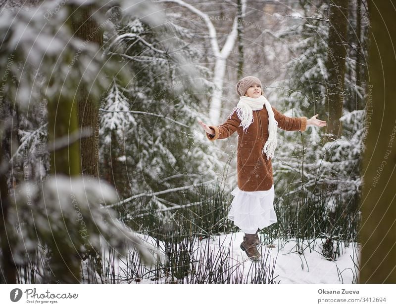 Ein 7-jähriges Mädchen in einem braunen Schafsfellmantel geht im Wald spazieren. Konzept der Jahreszeiten, ein Wintermärchen. Schnee, Bäume. Baby Hintergrund