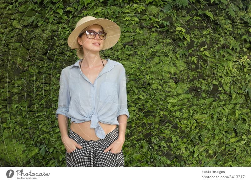 Porträt einer schönen Reisenden. Lächelnde junge Frau mit Sommerhut und Sonnenbrille, die vor einem üppigen tropischen Pflanzengrün vor dem Hintergrund der Wand steht.