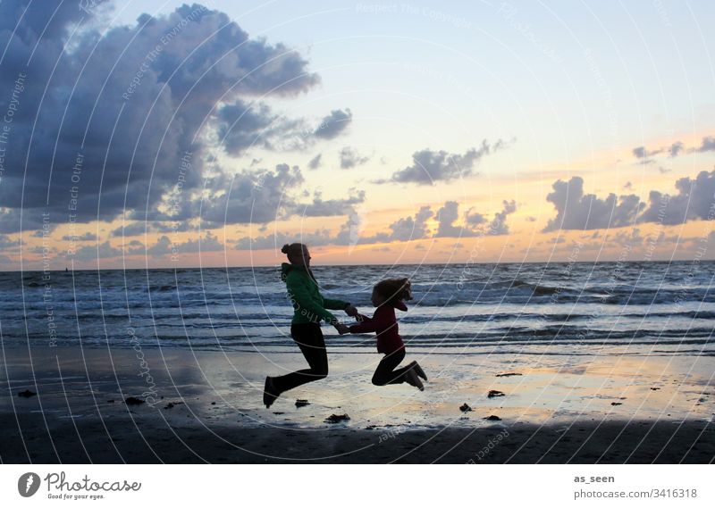 Zwei Mädchen machen einen Luftsprung am Meer im Sonnenuntergang Kontrast Silhouette Kind Kindheit Strand Wolken Sprung springen Spaß Freiheit Freude