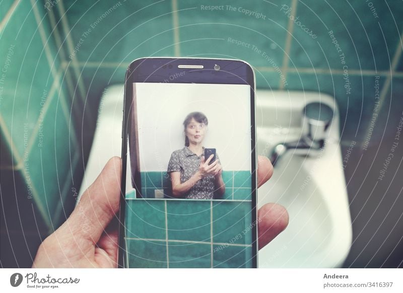 Vor einem Waschbecken in einem türkis gefliesten Bad hält eine linke Hand ein Handy mit einem Selfie einer Frau Smartphone Foto Fliesen Finger waschen