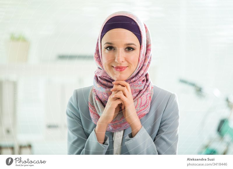Schöne, stilvolle Frau mit Hijab und Brille, die im Büro am Schreibtisch mit Laptop sitzt. Porträt einer selbstbewussten muslimischen Geschäftsfrau. Modernes Büro mit großem Fenster, Fahrrad im Hintergrund.