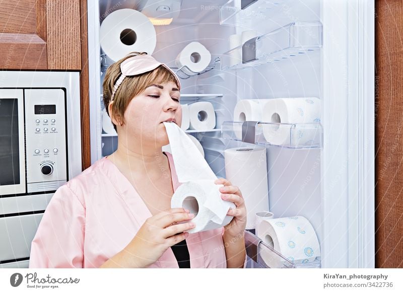 Frau im Schlafanzug isst gierig Toilettenpapier in der Nähe des Kühlschranks, das mit WC-Papier gefüllt ist Nacht Verlangen Coronavirus covid-19 Haushalt