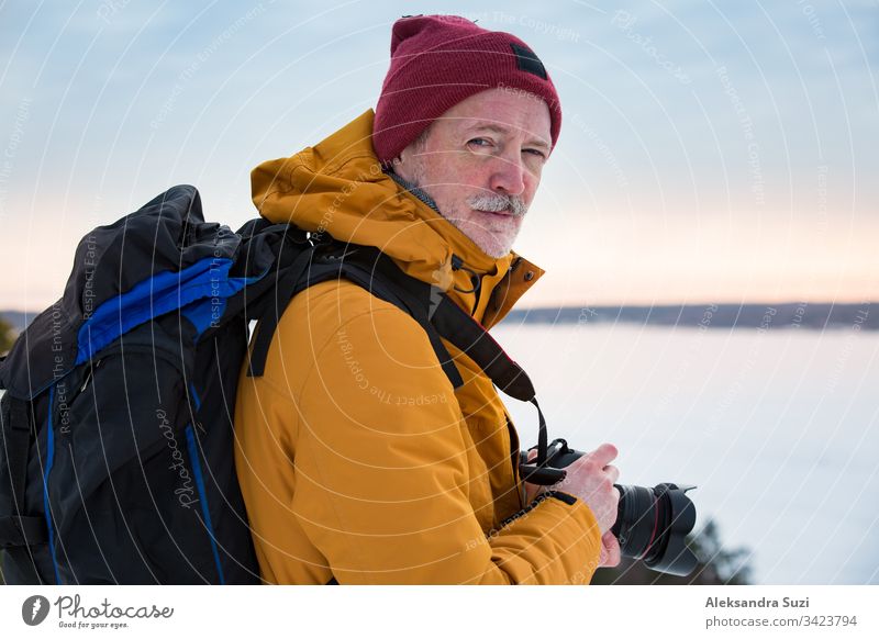 Porträt eines reifen Mannes mit grauem Bart, der im Winter Finnland erkundet. Reisender beim Fotografieren auf dem Gipfel eines Felsens. Schöne Aussicht auf die nördliche Landschaft mit gefrorener Ostsee und verschneiten Inseln.