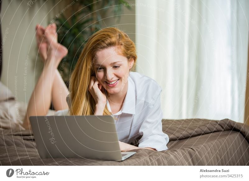 Junge Frau mit langen Haaren, die morgens im Schlafzimmer auf dem Bett liegt, ein Hemd trägt, online chattet, telefoniert, lächelt. Erwachsener schön Schönheit
