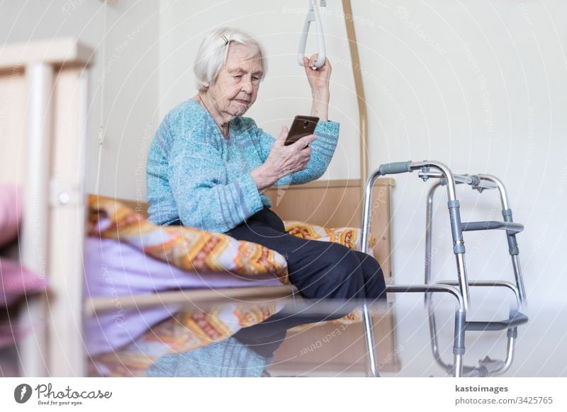 Ältere 96 Jahre alte Frau, die eine Telefonnachricht liest, während sie auf einem medizinischen Bett sitzt und von einem Halter gestützt wird. Senior heimwärts