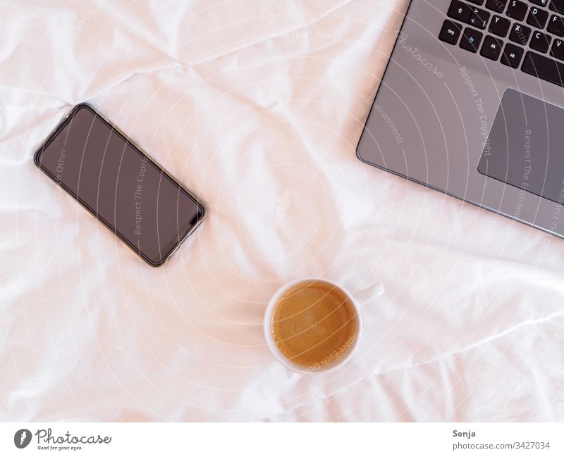 Laptop, Smartphone und eine Tasse Kaffee auf einer weißen Bettdecke, Heimbüro Heimarbeitsplatz Technik & Technologie Lifestyle Computer Business Internet