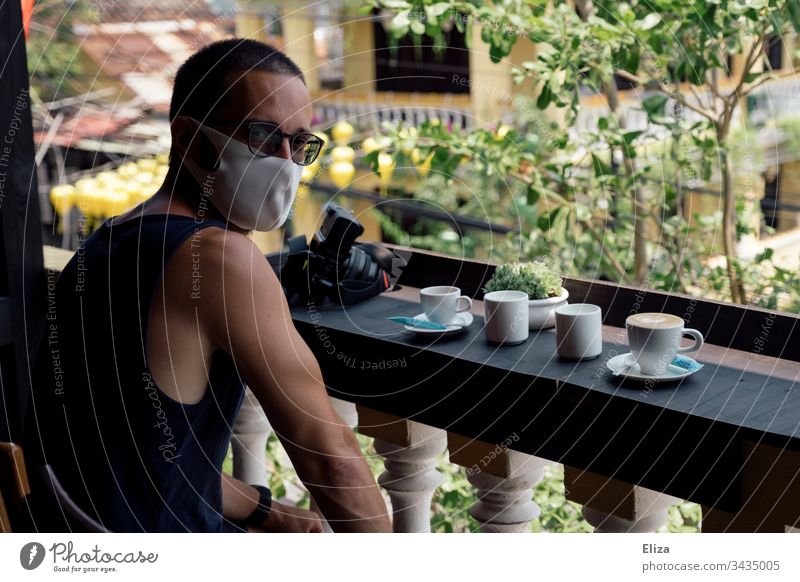 Ein Mann mit Gesichtsmaske sitzt in einem Café im Ausland, wo er wegen des Coronavirus gestrandet ist Covid Covid-19 Tourist Leben Urlaub Asien Kaffee Sonne