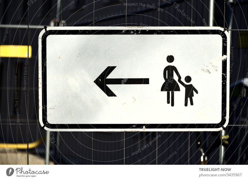 An einem Absperr-Gitter ein weisses Hinweis-Schild für Fußgänger mit schwarzem Piktogramm, das einer stilisierten Frau mit kleinem Kind an der Hand durch einen Pfeil signalisiert: Weitergehen nach Links. | Corona Thoughts