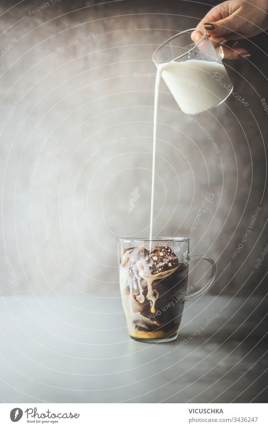 Zubereitung von Eiskaffee. Weibliche Hand gießt Sahne-Milch in Glas mit Kaffee-Eiswürfeln auf rustikalem Tisch im Betonwandhintergrund. Zubereitung von Eiskaffee. Erfrischendes Sommergetränk. Kaltes Getränk.