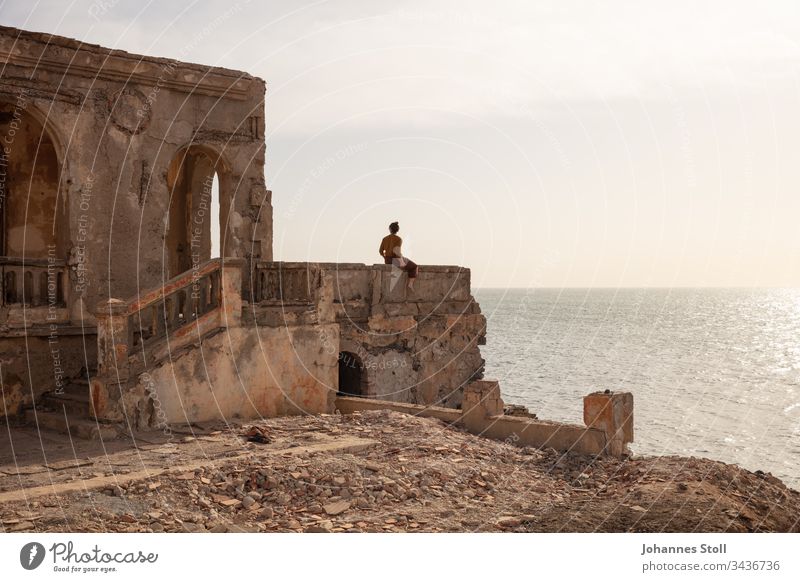 Junge Frau sitzt auf Mauerbrüstung einer Ruine und blickt aufs Meer Ozean Wasser Strand Küste Trümmer Steine Fels Mauerwerk Architektur Tempel Burg Schloss