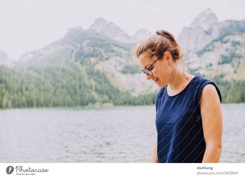 junge Frau schaut lächelnd auf dem Boden vor einem See mit Gipfeln im Hintergrund Jugendliche leuchtend attraktiv 1 Mensch Erwachsene brünett wandern Tourismus
