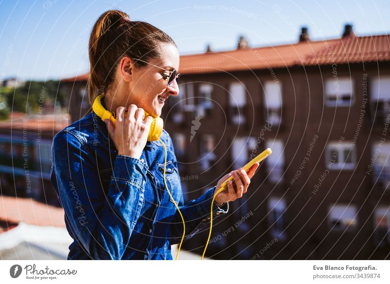 junge Frau, die über ein Mobiltelefon und ein gelbes Headset Musik hört. Spaß und Lebensstil hören Handy Technik & Technologie Terrasse im Freien sonnig