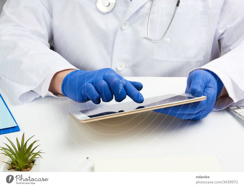 Arzt im weißen Kittel sitzt an einem Tisch, hält ein elektronisches Tablett in der Hand, berührt mit der rechten Hand den Bildschirm Tablette medizinisch