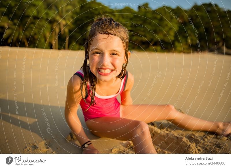 Süßes fröhliches kleines Mädchen, das im Badeanzug am Strand mit Sand spielt, ein Herz zeichnet und schreibt. Wunderschöner Sommersonnenuntergang, Meer, Kokospalmen, malerische exotische Landschaft. Phuket, Thailand