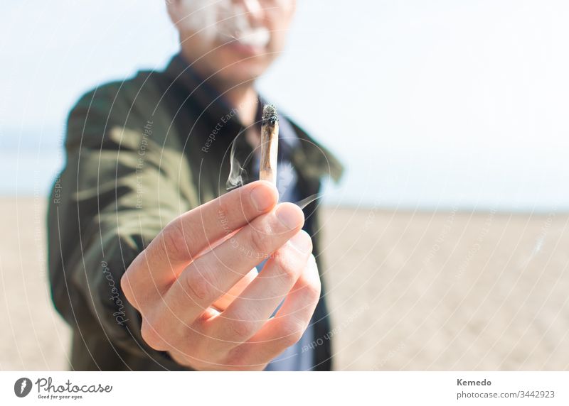 Junger Mann hält einen angezündeten Marihuana-Joint in der Hand, während er am Strand raucht. Hintergrund und Kopierraum rechts verwischen. Gelenk Person