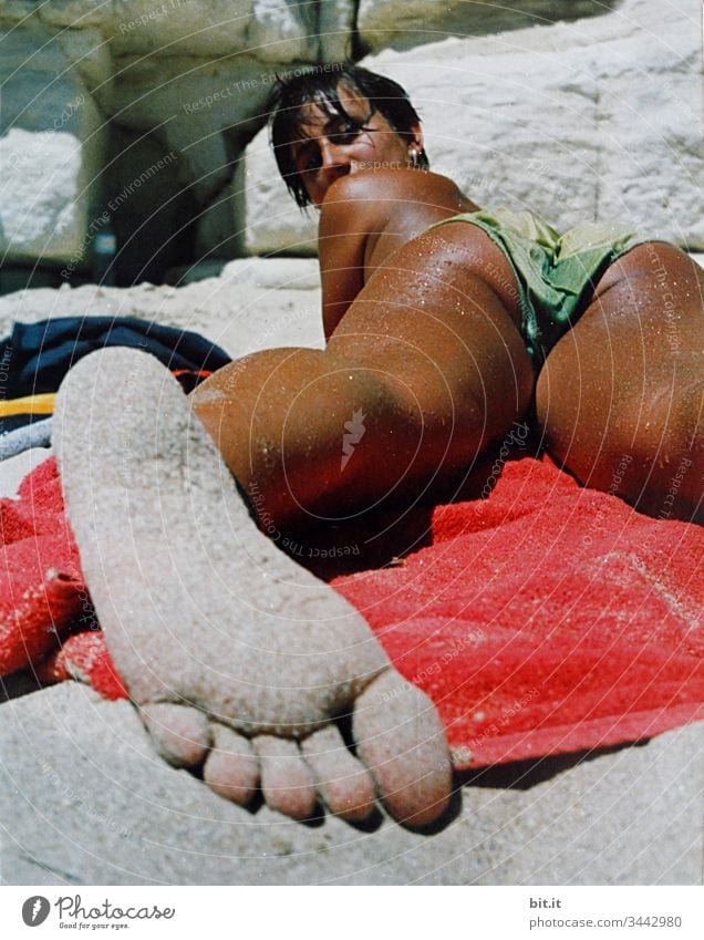 Junge Frau liegt braungebrannt am Strand auf einem Handtuch, barfuß mit sandigem Fuß im Vordergrund und schaut in die Kamera. Sonnenbad Sommer