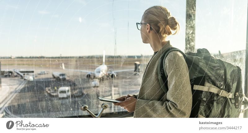 Junge Frau steht in der Nähe des Flughafenfensters, hält ihr Handy in der Hand, trägt einen Reiserucksack und geht zu Fuß zum Lounge-Bereich. Reisende Frau sucht Online-Karte am Flughafen