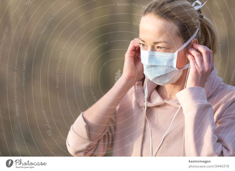Porträt einer kaukasischen, sportlichen Frau, die eine medizinische Schutzmaske trägt, während sie im Park spazieren geht, sich entspannt und Musik hört. Der Corona-Virus, oder Covid-19, verbreitet sich weltweit.
