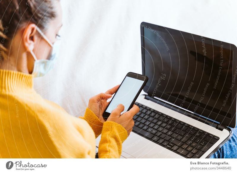 junge Frau, die zu Hause am Laptop arbeitet, auf der Couch sitzt und eine Schutzmaske trägt. Das Konzept "zu Hause bleiben" während des Coronavirus covid-2019