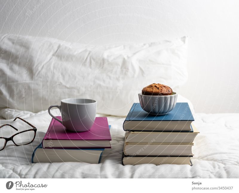 Bücherstapel mit einer Tasse Kaffee und einem Muffin auf einer weißen Bettdecke im Schlafzimmer bücherstapel Hygge bettwäsche weiss schlafzimmer vereinzelt