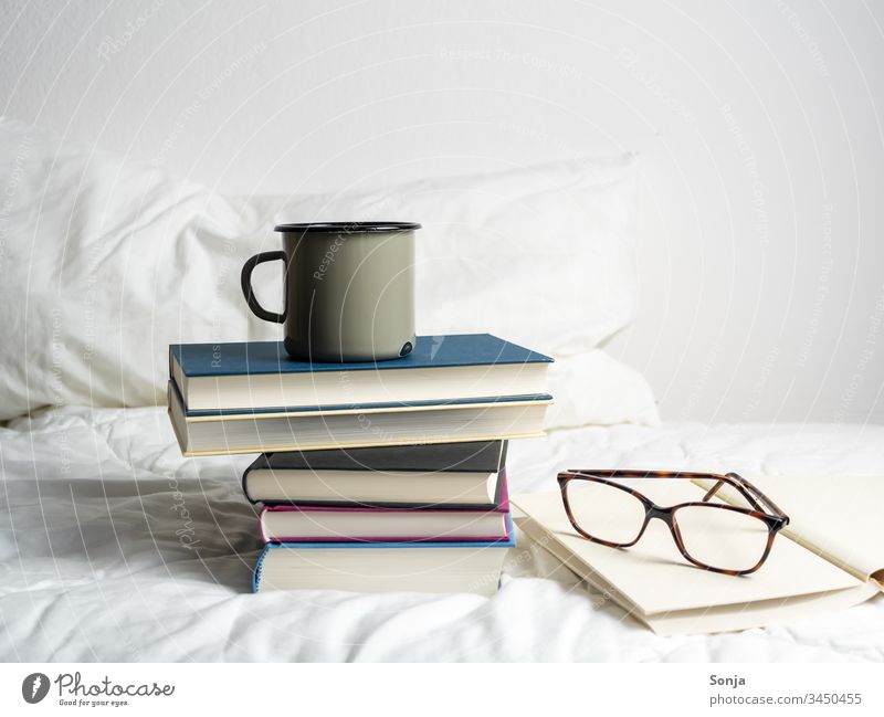 Bücherstapel mit einer Tasse Kaffee und einer Lesebrille auf einer weißen Bettdecke im Schlafzimmer bücherstapel getränk lesebrille bettdecke weiss schlafzimmer