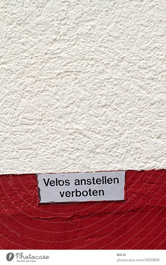 Schild an einer rot, weissen Hauswand aus Stein, in der Schweiz, mit Text: Velos anstellen verboten, als Hinweis auf Parkverbot für Fahrräder an der Mauer, in der Stadt.