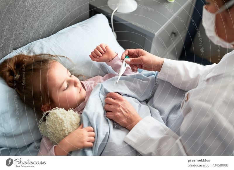 Der Arzt besucht den kleinen Patienten zu Hause. Messung der Temperatur eines kranken Mädchens, das im Bett liegt. Frau trägt Uniform und Gesichtsmaske. Medizinische Behandlung