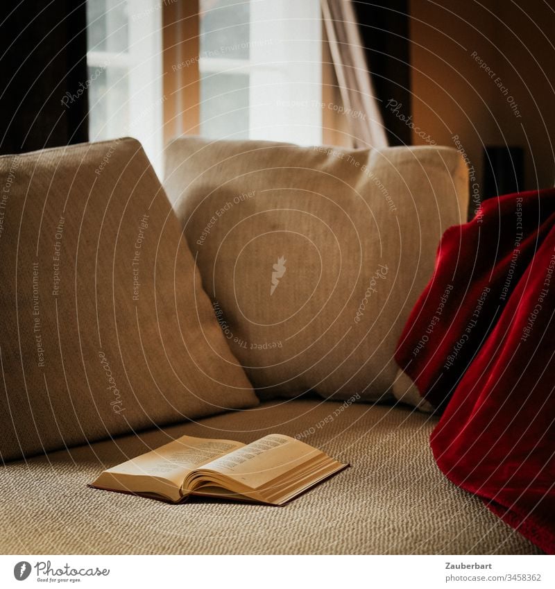 Gemütliches Sofa mit Kissen, roter Decke und Buch, im Hintergrund Sprossenfenster, als Leseplatz zuhause stayhome lesen gemütlich Fenster Tageslicht beige