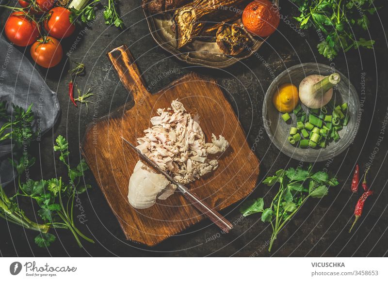 Gezogene Hühnerbrust auf Schneidebrett mit Messer mit Gemüsezutaten auf dunklem, rustikalem Hintergrund. Ansicht von oben. Gesunde Ernährung. Kohlenhydratarme Fitness-Diät. Leichtes Kochen