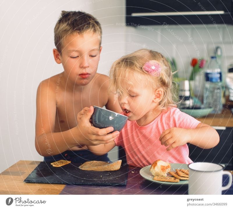 Der ältere Bruder kümmert sich um die jüngere Schwester und hilft ihr beim Trinken aus der Tasse während des Frühstücks. Alltägliche Morgenmomente Kinder Pflege